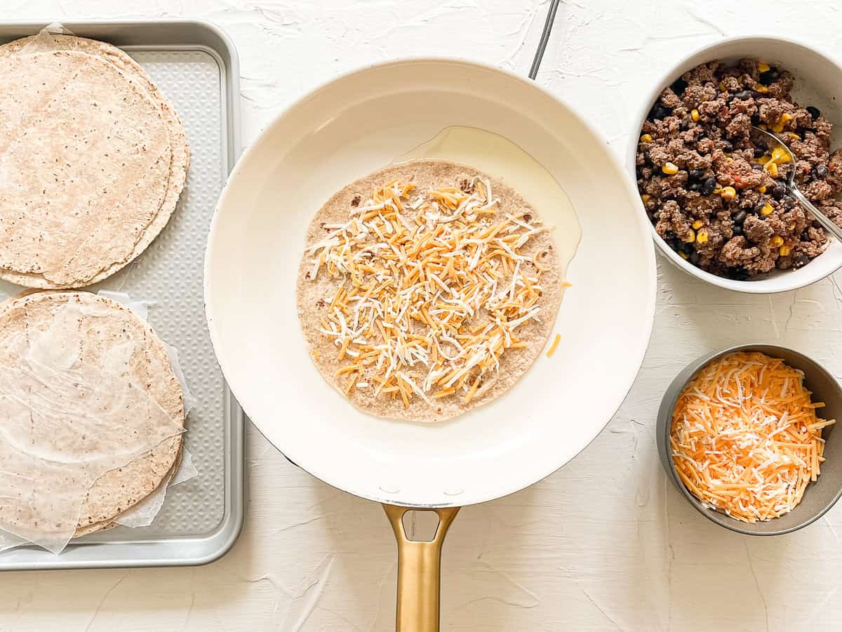 Quesadilla making station with a skillet in the middle with shredded cheese on a tortilla.