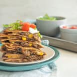 Quesadilla servings stacked up on a plate with sour cream, chopped tomatoes, and shredded lettuce on top.