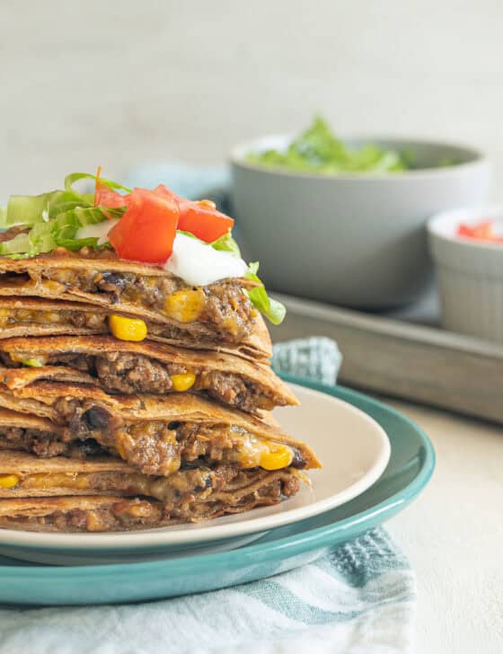 Quesadilla servings stacked up on a plate with sour cream, chopped tomatoes, and shredded lettuce on top.