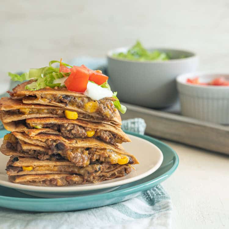 Quesadilla servings stacked up on a plate with sour cream, chopped tomatoes, and shredded lettuce on top.