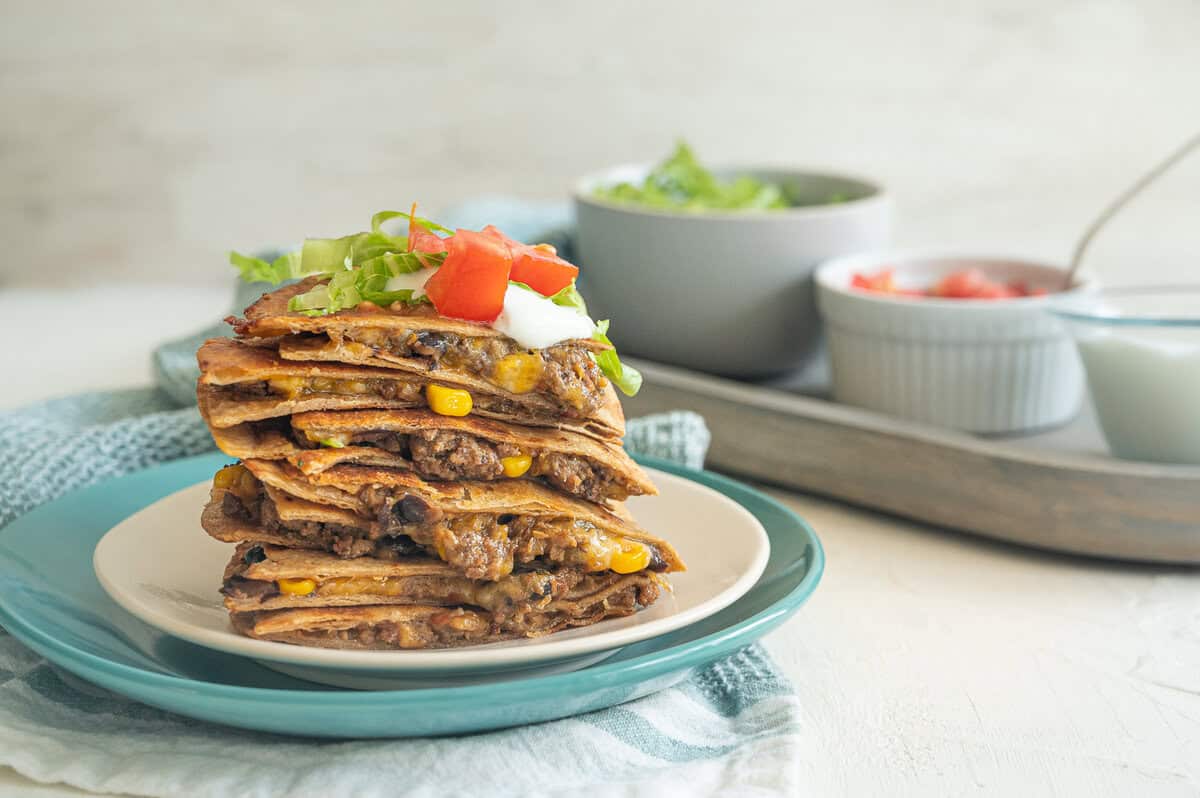 Quesadilla servings stacked up on a plate with sour cream, chopped tomatoes, and shredded lettuce on top.