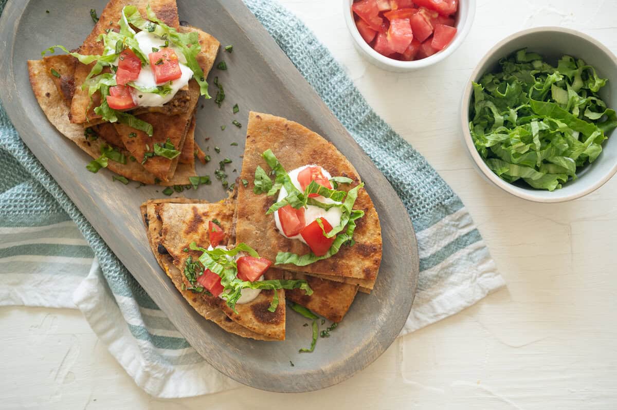 A quesadilla cut in quarters, stacked on a serving platter with sour cream, chopped tomatoes, and shredded lettuce.