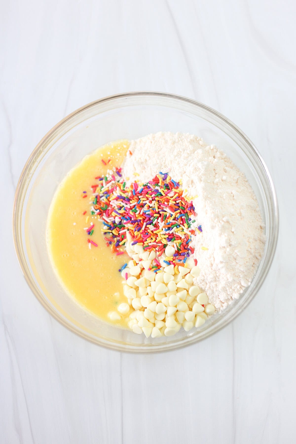 Cake mix, sprinkles, and white chocolate chips on top of wet ingredients for Funfetti cake mix cookies in a mixing bowl.