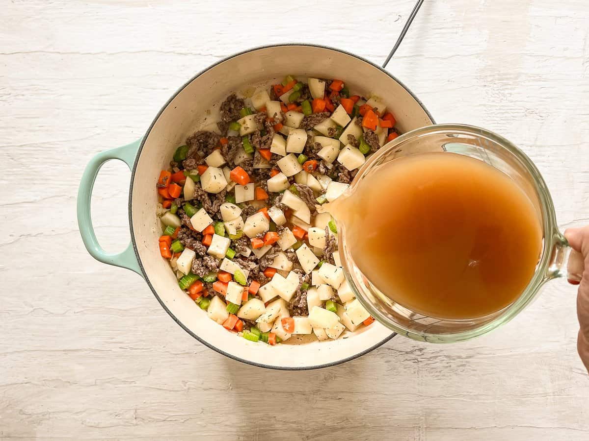 Broth being poured into a ceramic pot that has sauteed veggies, chopped potatoes, browned ground beef, and seasonings in it.