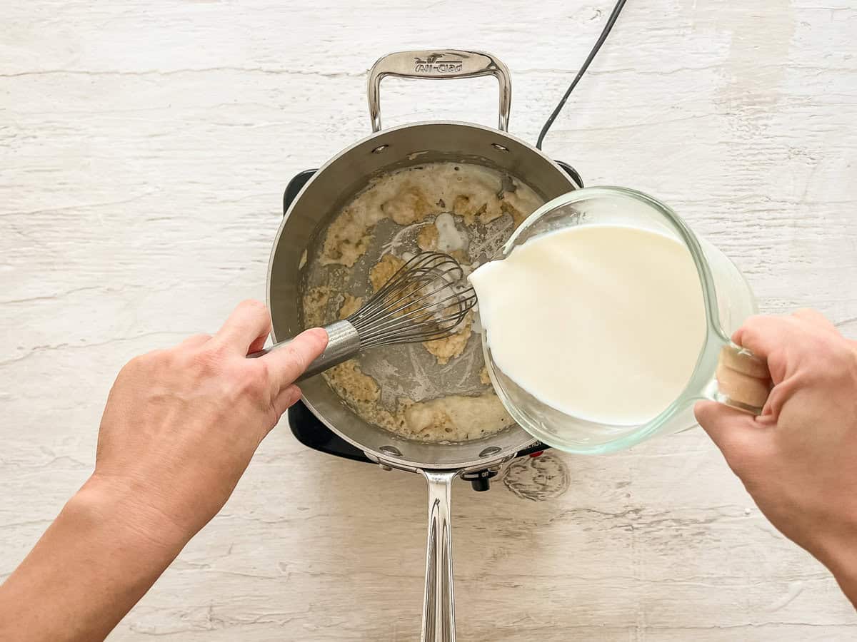 Flour and butter being whisked together while milk is being poured into a sauce pan for a roux.