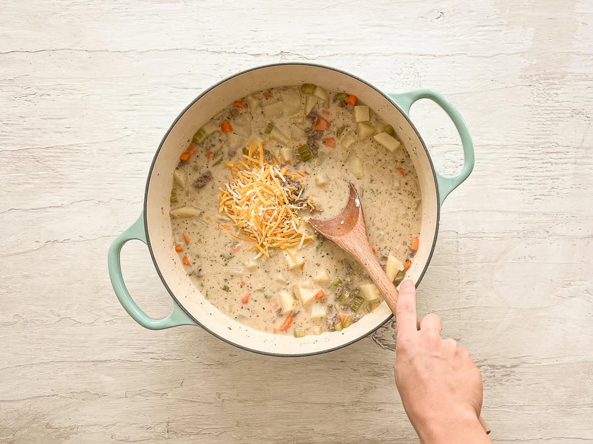 Shredded cheese being mixed into a ceramic pot with ingredients for cheeseburger soup.