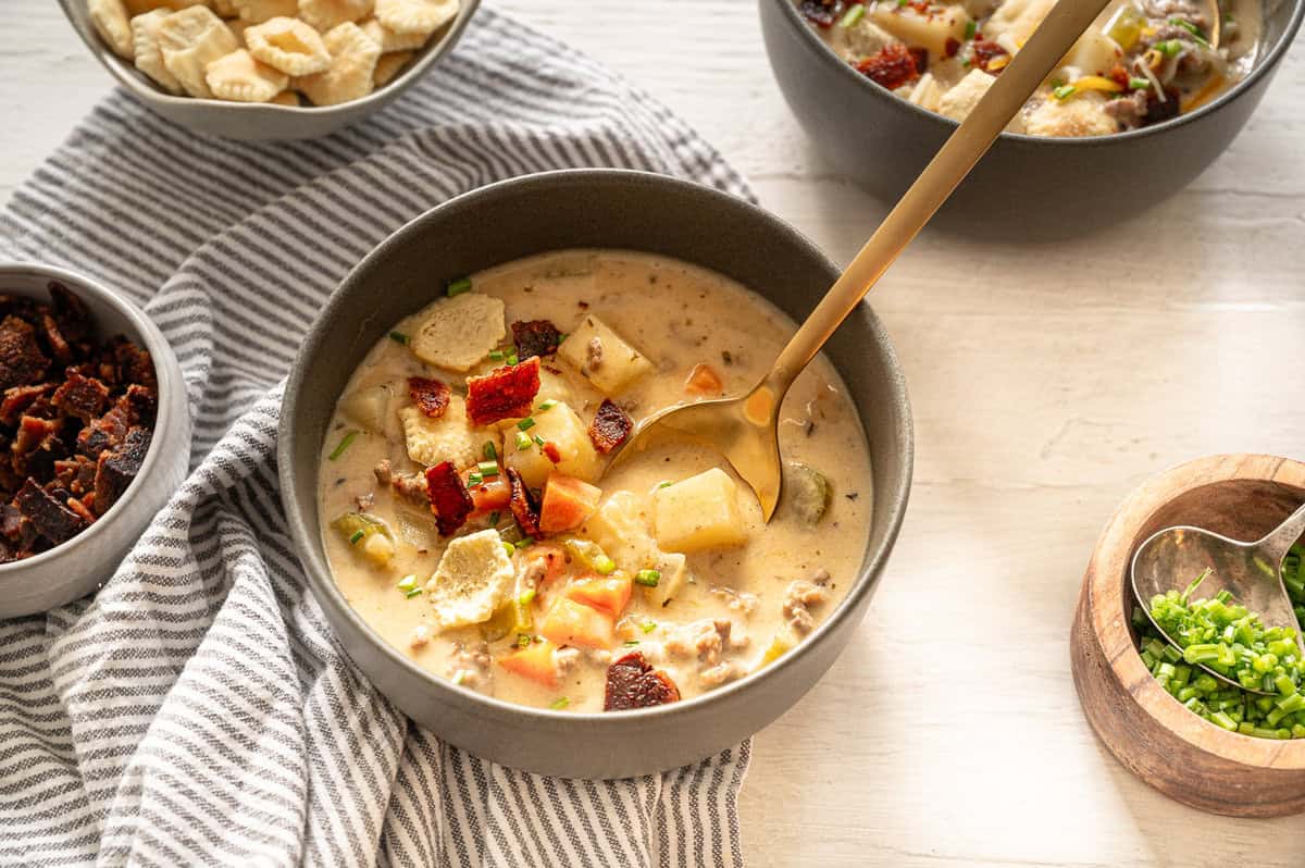 A bowl of cheeseburger soup with small bowls of chopped green onions and bacon next to it.