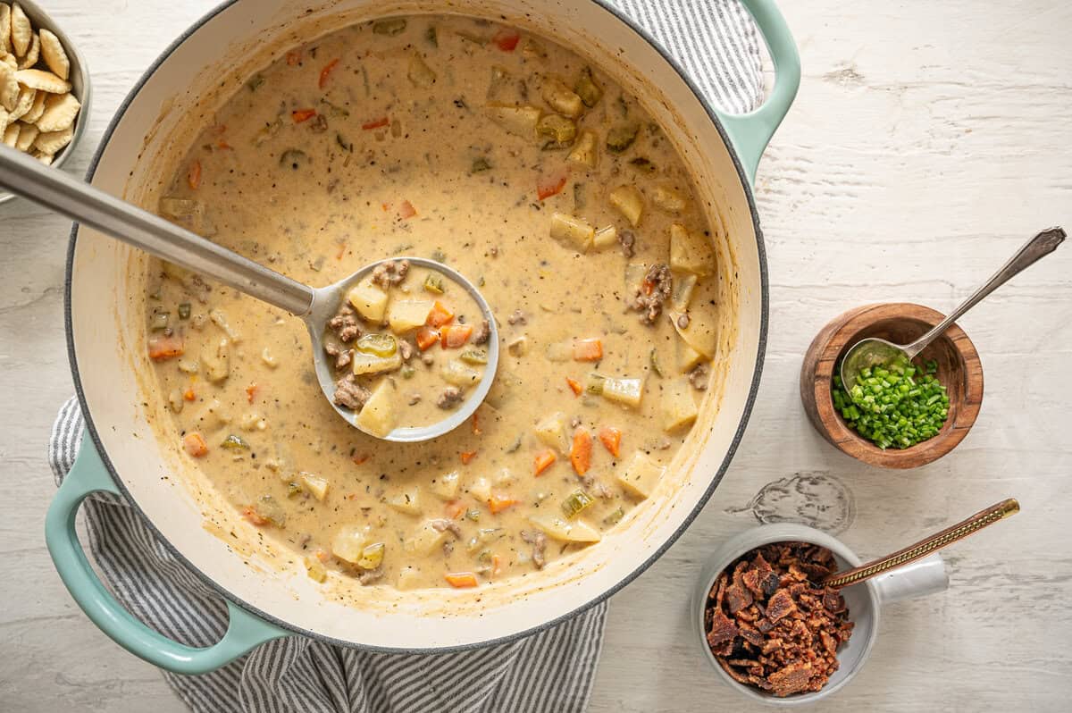 A ladle of cheeseburger soup being lifted out of a dutch oven with small bowls of chopped green onions, and bacon next to it.