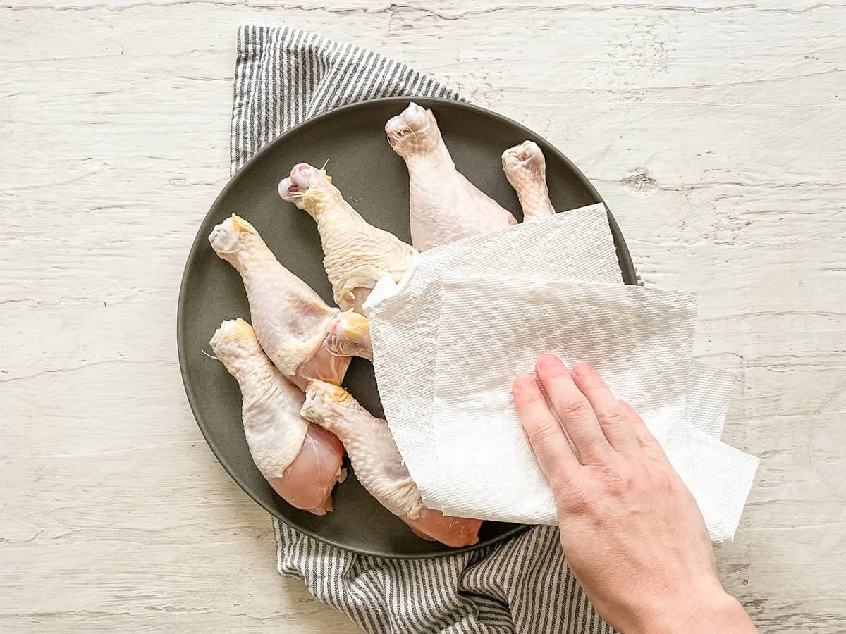 A hand blotting chicken drumsticks with a paper towel.