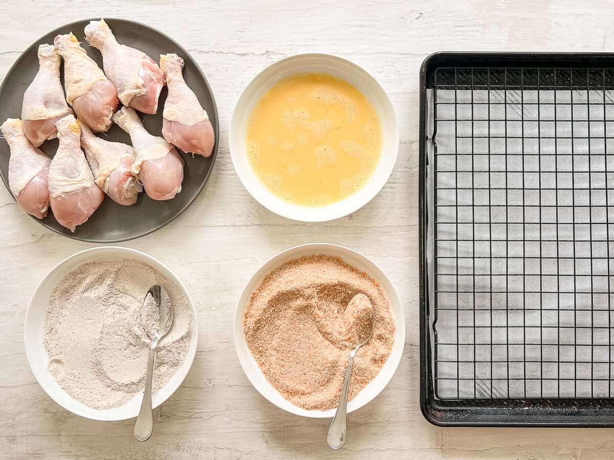 Crispy baked drumstick coating station set up with plates of drumsticks, beaten eggs, flour, and bread crumbs, and then a baking rack on a parchment lined baking sheet.