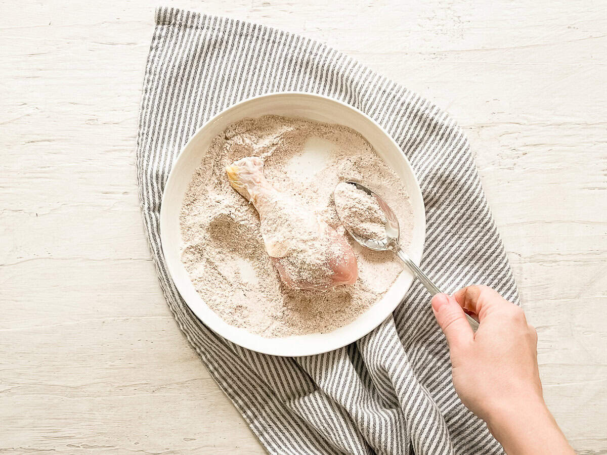 A hand using a spoon to coat a chicken drumstick with flour and spices on a plate.