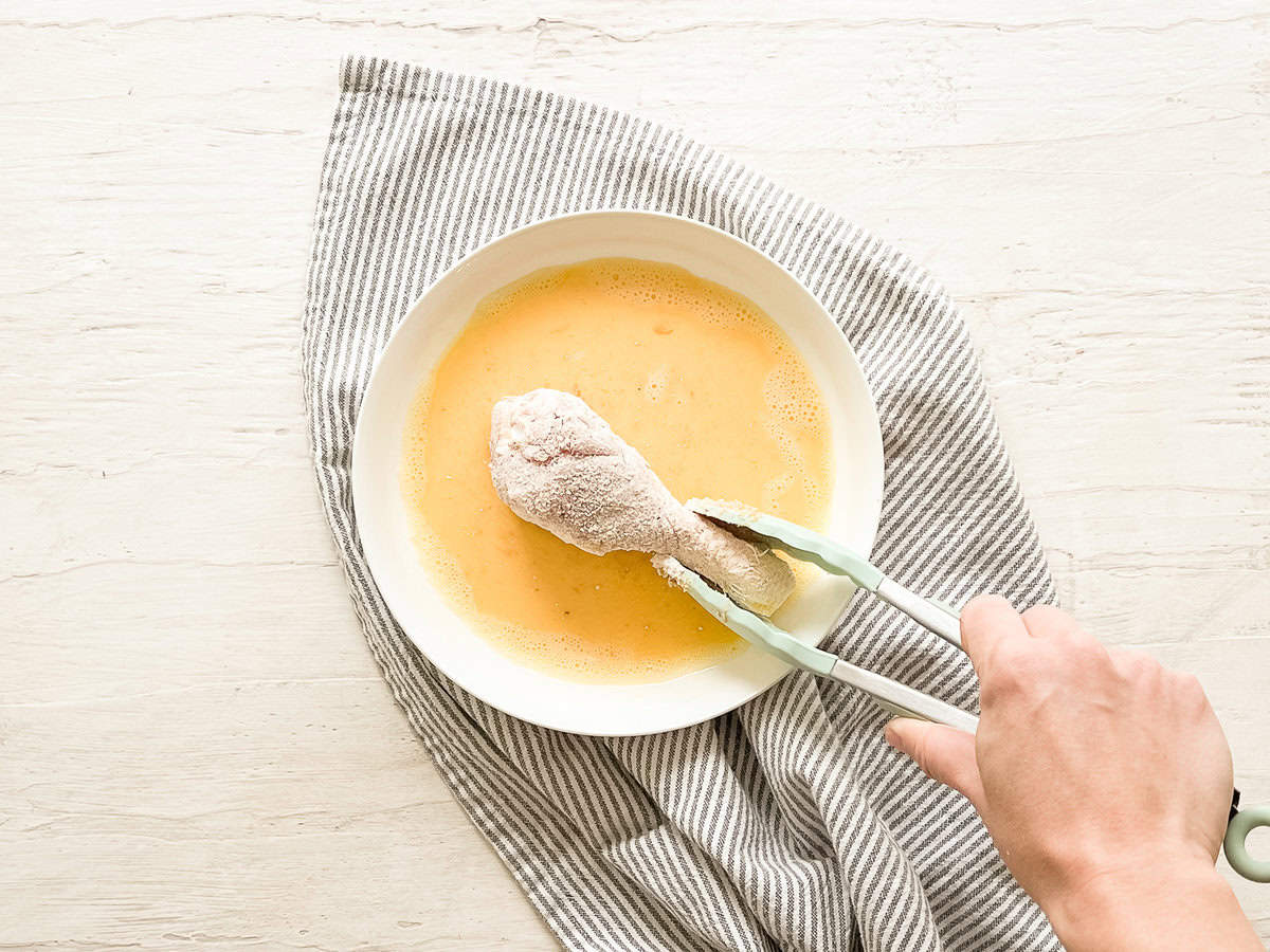 A hand using tongs to dip a flour-coated drumstick into a bowl with beaten eggs.