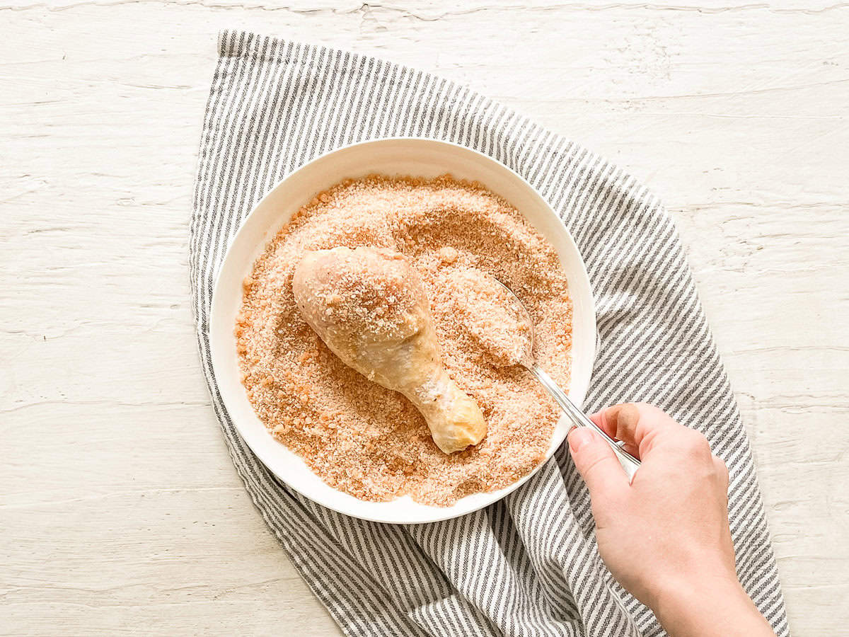 A hand using a spoon to cover a drumstick that was already coated in flour and beaten eggs, with breadcrumbs.