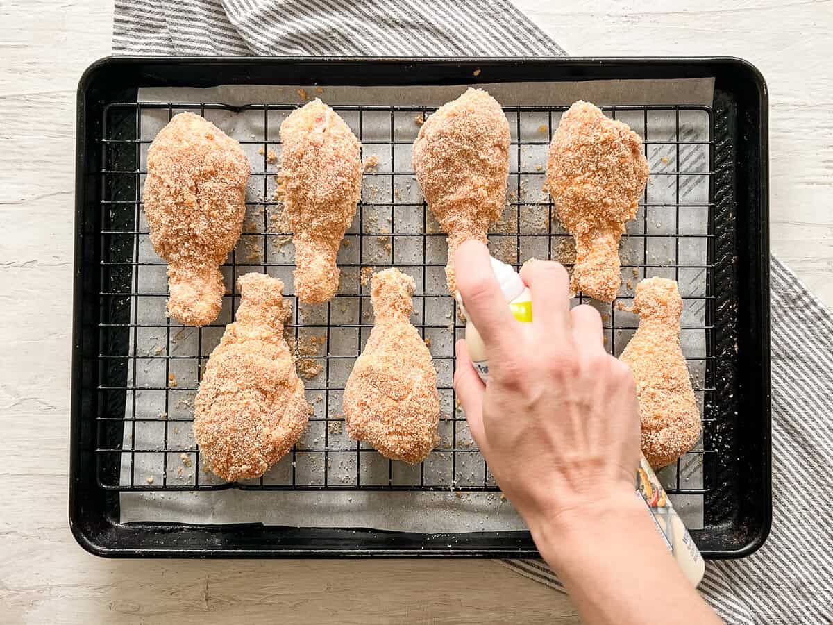 A hand spraying cooking oil onto breaded chicken drumsticks on a parchment-lined baking tray with rack.