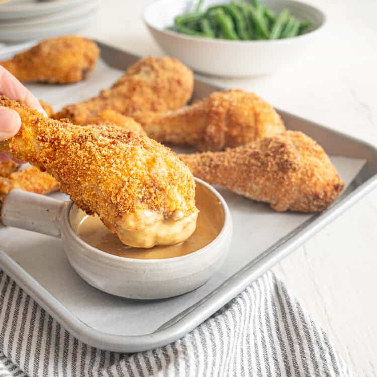 A hand dipping a crispy baked drumstick into a bowl of honey mustard sauce.