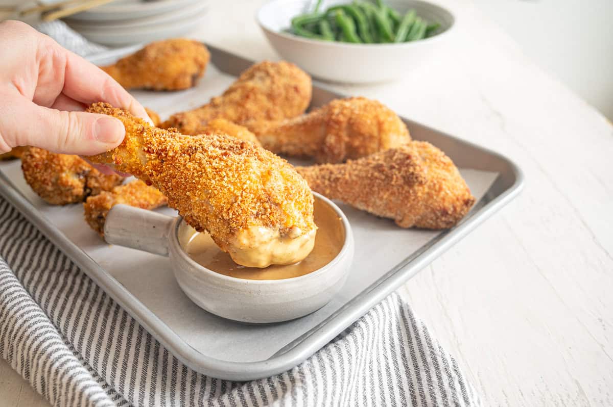 A hand dipping a crispy baked drumstick into a bowl of honey mustard sauce.