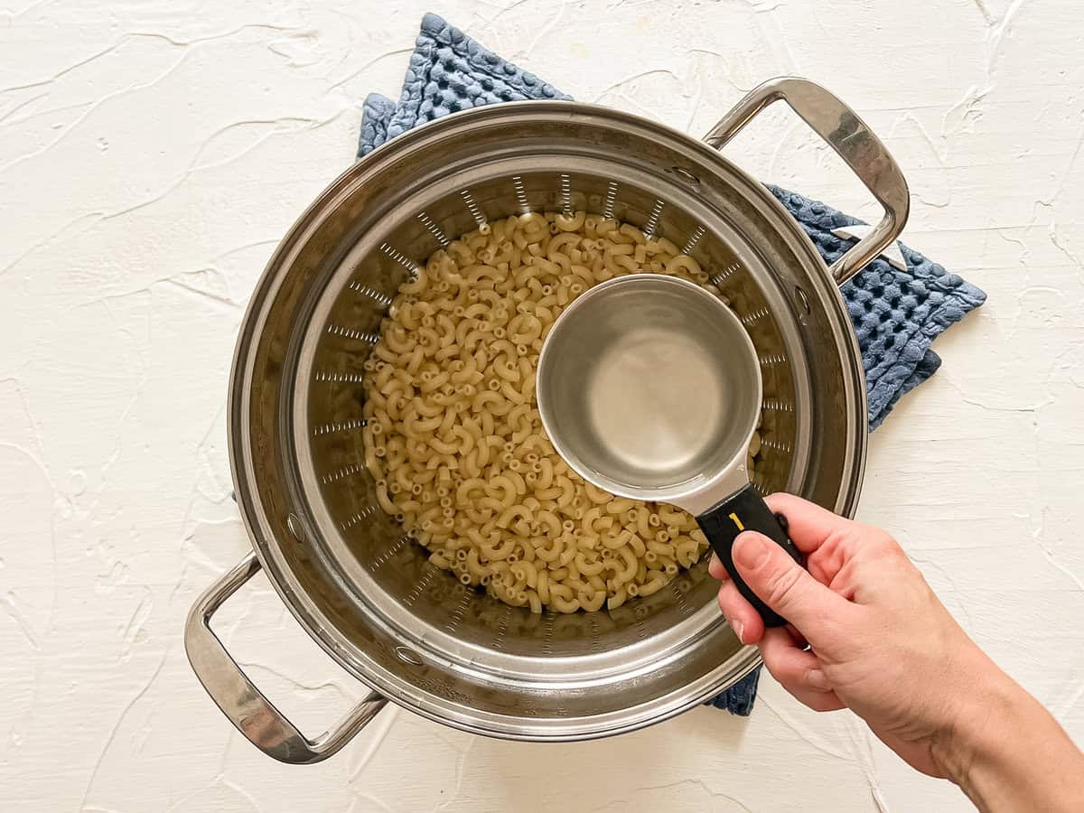 A hand removing a cup of pasta water before draining the rest of the liquid in a metal colander.