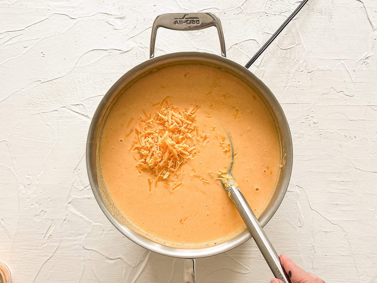 Shredded cheddar cheese being mixed into a sauce pan for homemade mac and cheese.