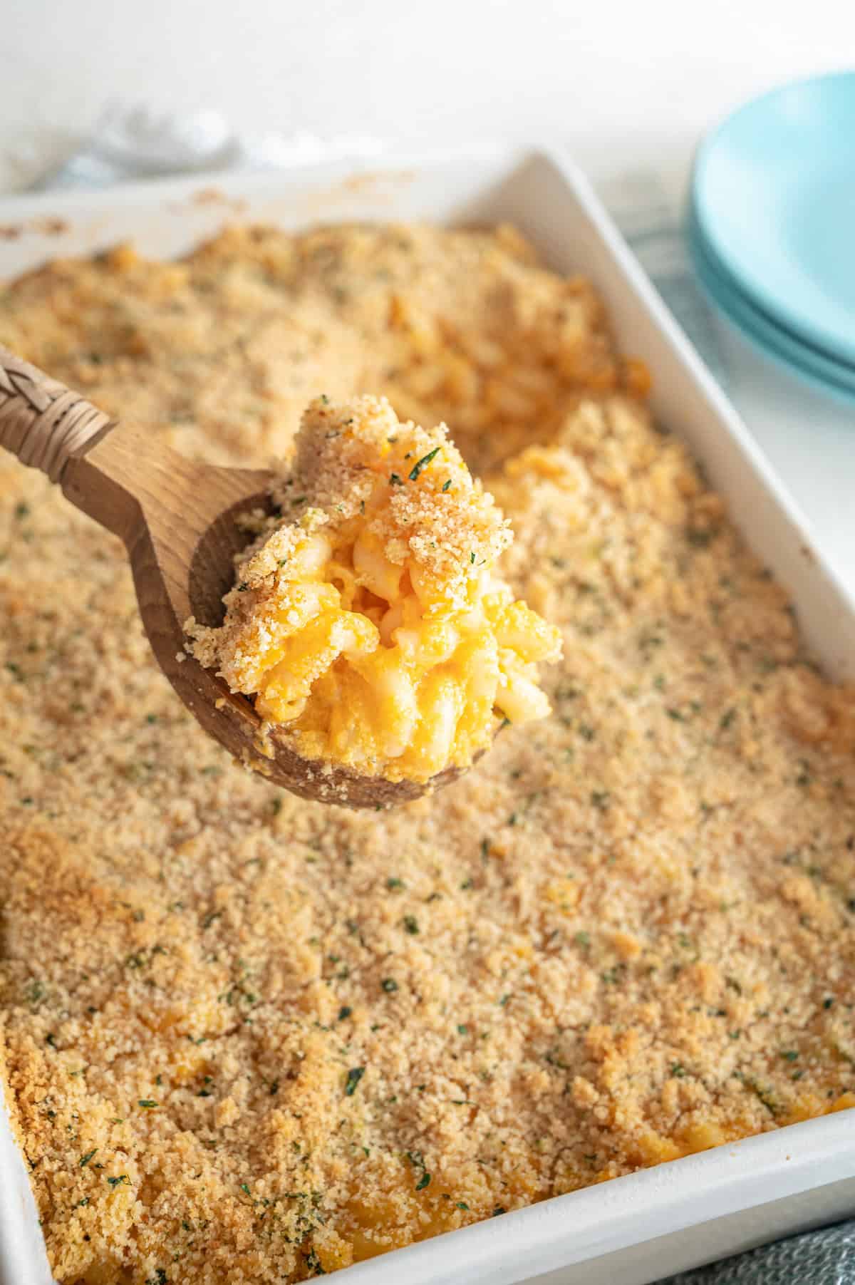 A wooden spoon lifting a scoop of homemade baked mac and cheese from a 9x12 white baking dish.