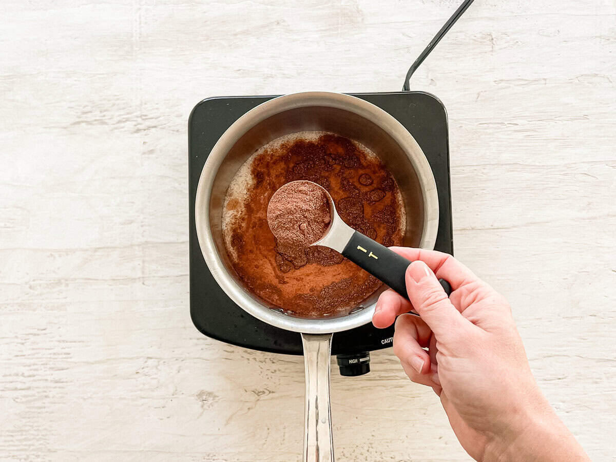 A hand holding a tablespoon of homemade hot chocolate mix over a pan of milk with more hot chocolate mix in it.