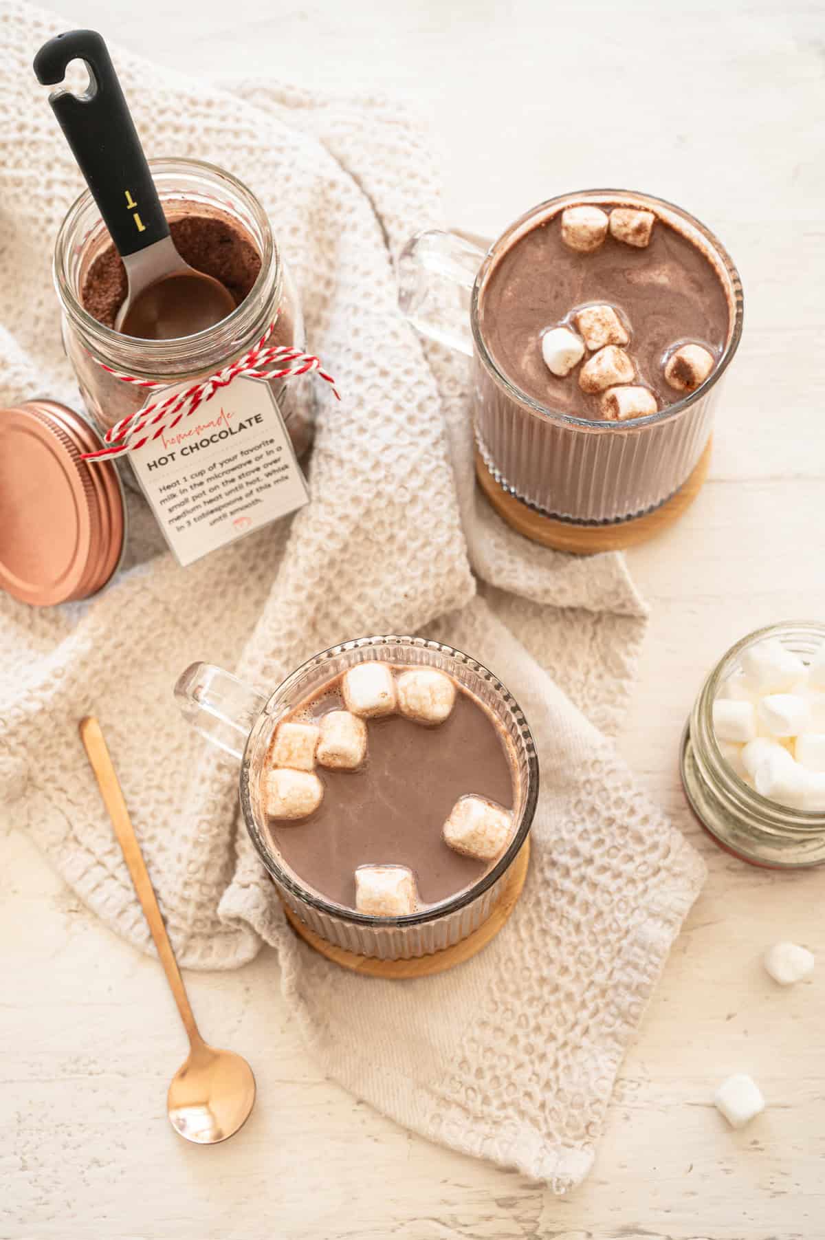 A mason jar of homemade hoc chocolate mix with two glass mugs of hot chocolate ready to drink.