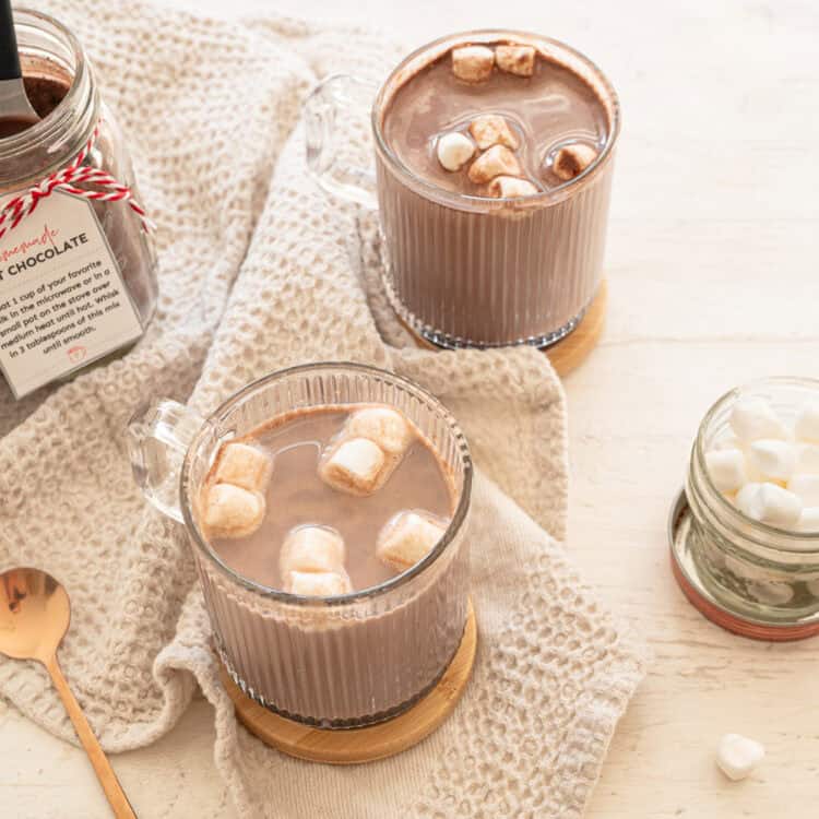 Homemade hot chocolate mix in a mason jar with two mugs of hot chocolate and mini marshmallows ready to drink.