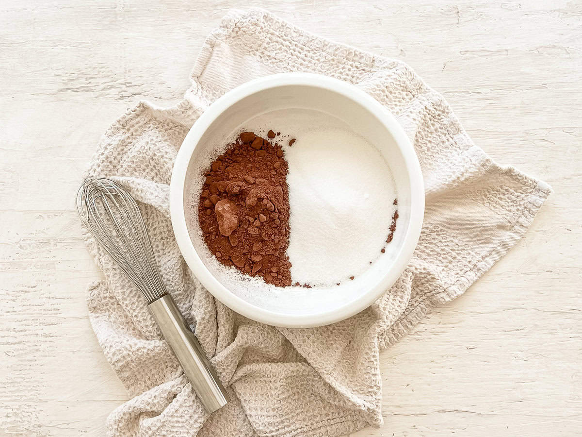 A small bowl with unsweetened cocoa powder on one side and sugar on the other side with a whisk next to it.