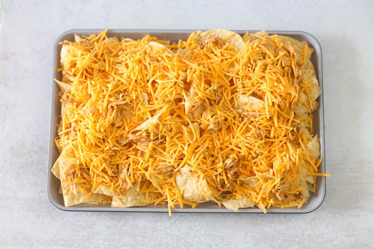 Tortilla chips covered in shredded cheese prepped for the oven on a baking sheet. 