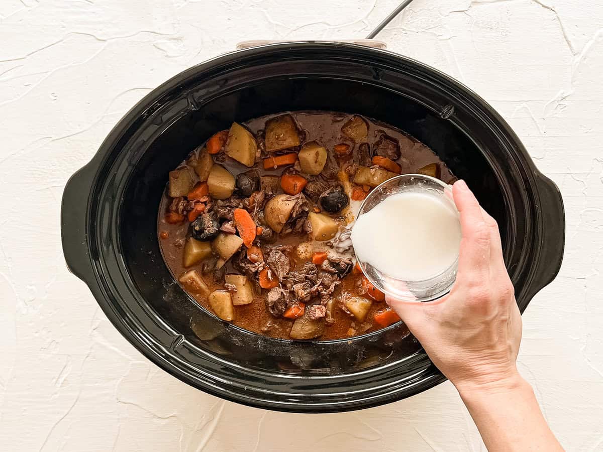 A hand pouring a cornstarch slurry into a crockpot of beef bourguignon.