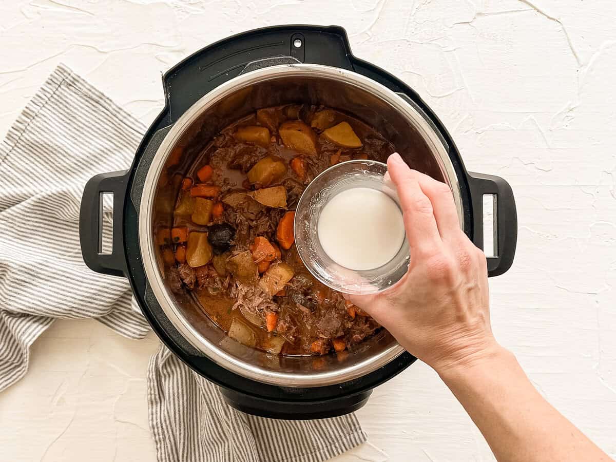 A hand pouring a cornstarch slurry into an Instant Pot of beef bourguignon.