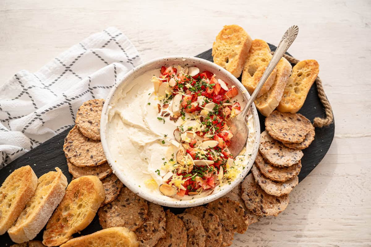 Honey whipped goat cheese in a bowl with chopped strawberries, sliced almonds, lemon zest, and fresh mint on a tray with crackers.