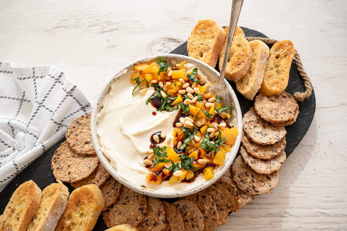 Honey whipped goat cheese with diced fresh peaches, pine nuts, fresh basil, and a drizzle of balsamic reduction in a ball on a tray of crackers.