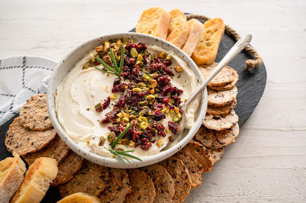 A tray of crackers and a bowl of honey whipped goat cheese with chopped pistachios, dried cranberries, and rosemary.