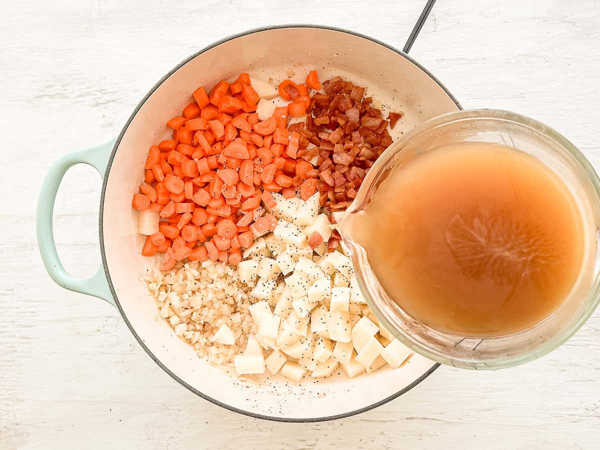 Chopped carrots, potatoes, and cooked bacon in a pot with chicken broth being poured into it.