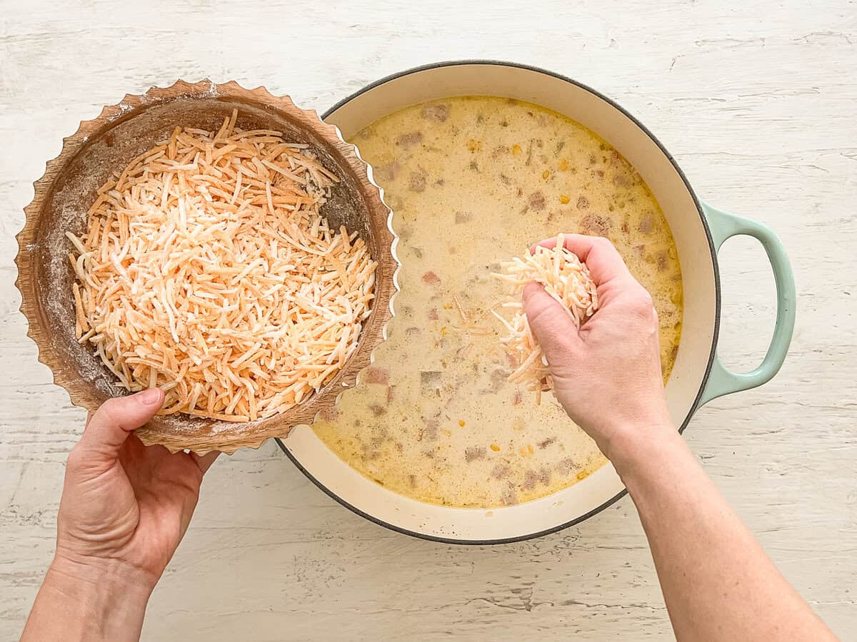 Flour-covered shredded cheddar cheese being placed by the handful into a pot of autumn chowder.