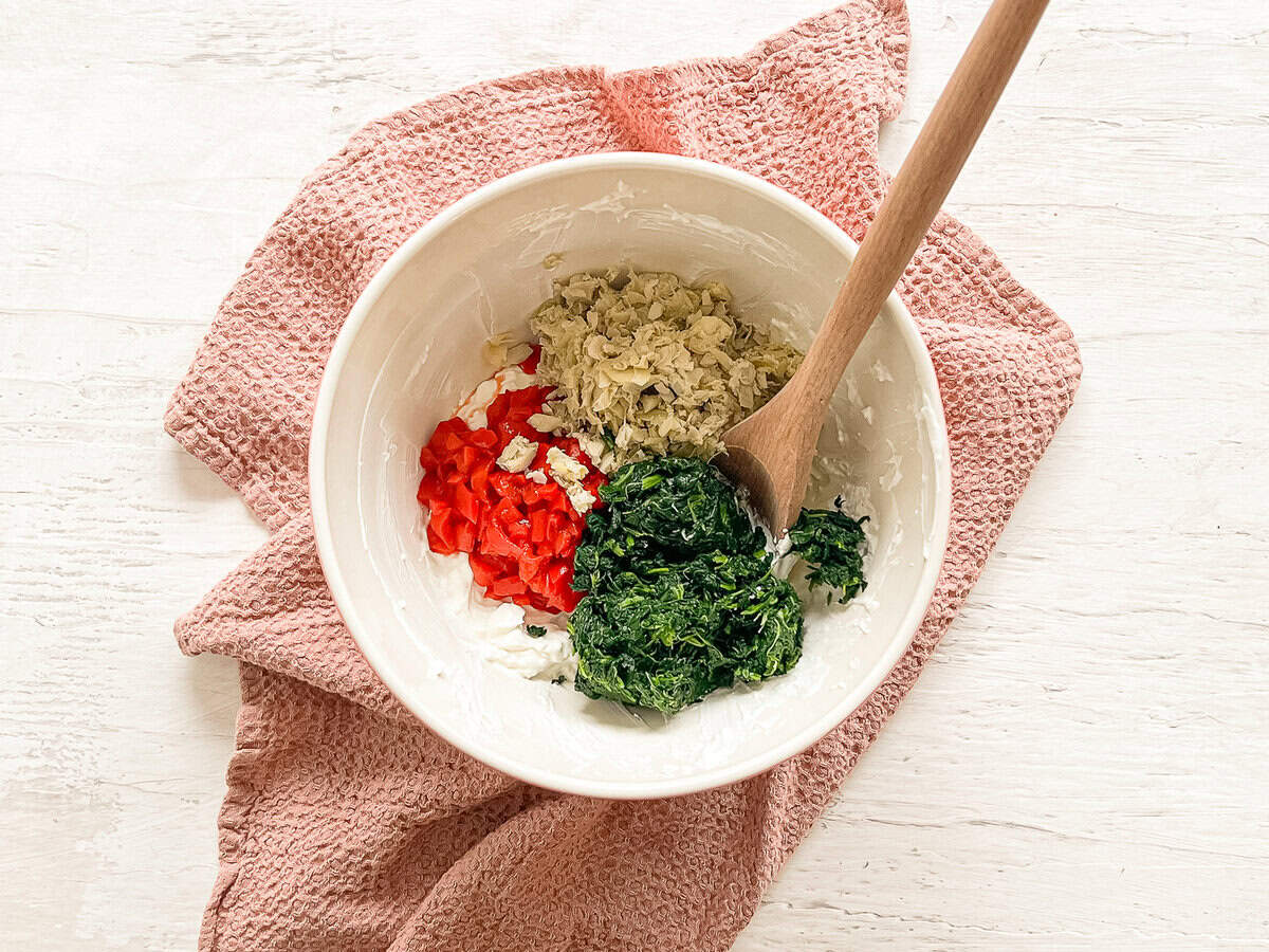 A bowl with piles of frozen chopped spinach, chopped roasted red peppers, and chopped artichoke hearts on a cream cheese base in a bowl ready to be mixed together.