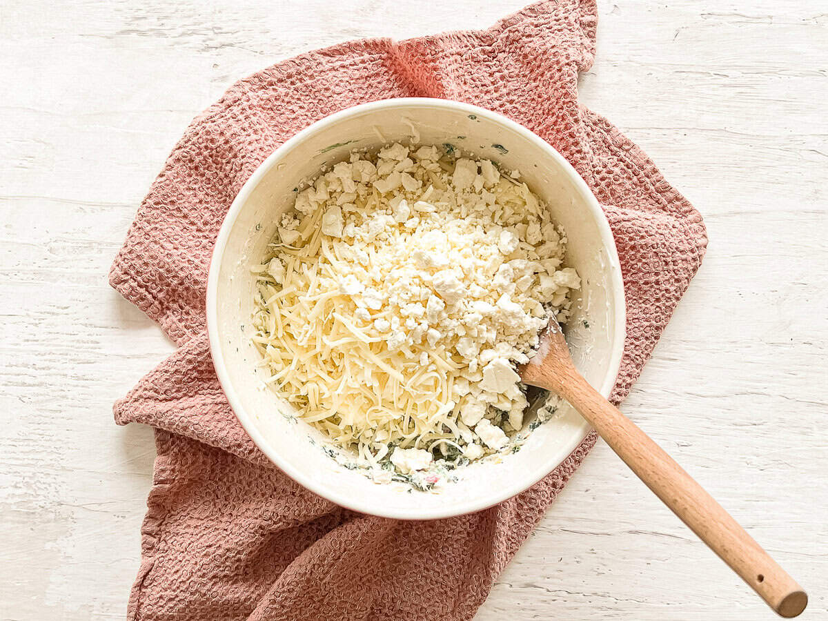 A cream cheese base of spinach artichoke dip in a bowl with mozarella and feta cheeses on top ready to be mixed in.