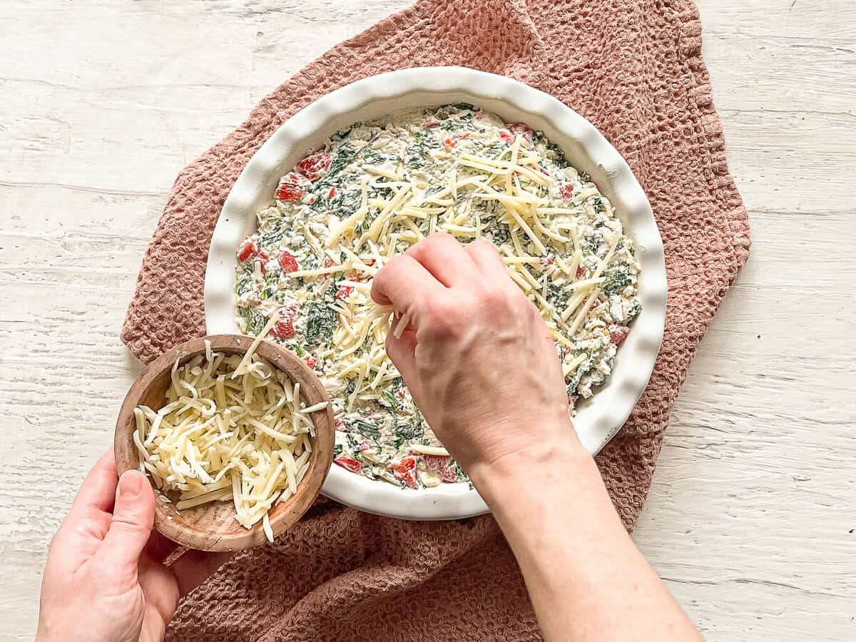 A hand sprinkling mozarella cheese on top of a crock with spinach and artichoke dip in it.