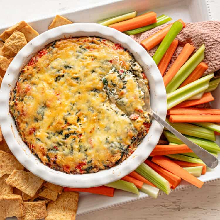 Crock of baked spinach and artichoke dip on a platter of carrots, celery, and crackers.