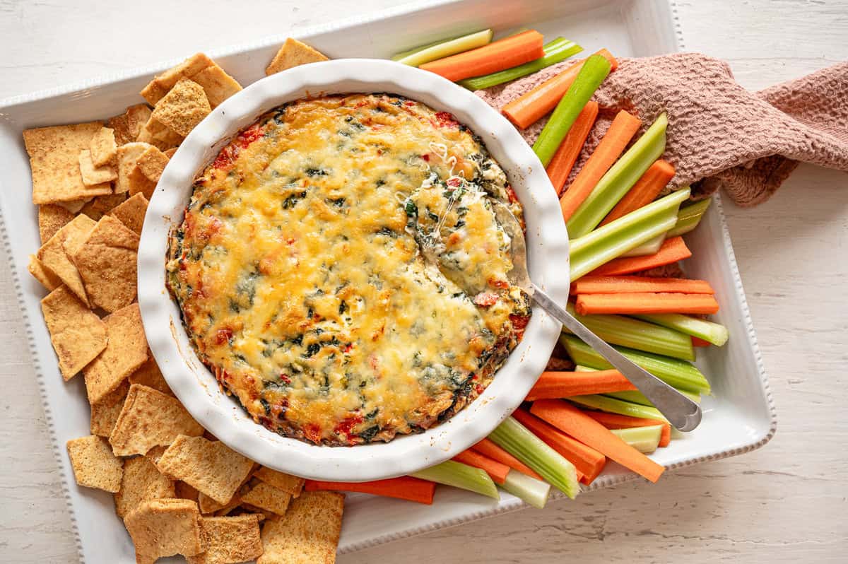 Crock of baked spinach and artichoke dip on a platter of carrots, celery, and crackers.