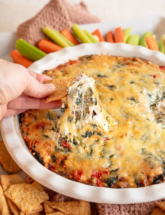 A cracker lifting some spinach and artichoke dip out of the crock it was made in.