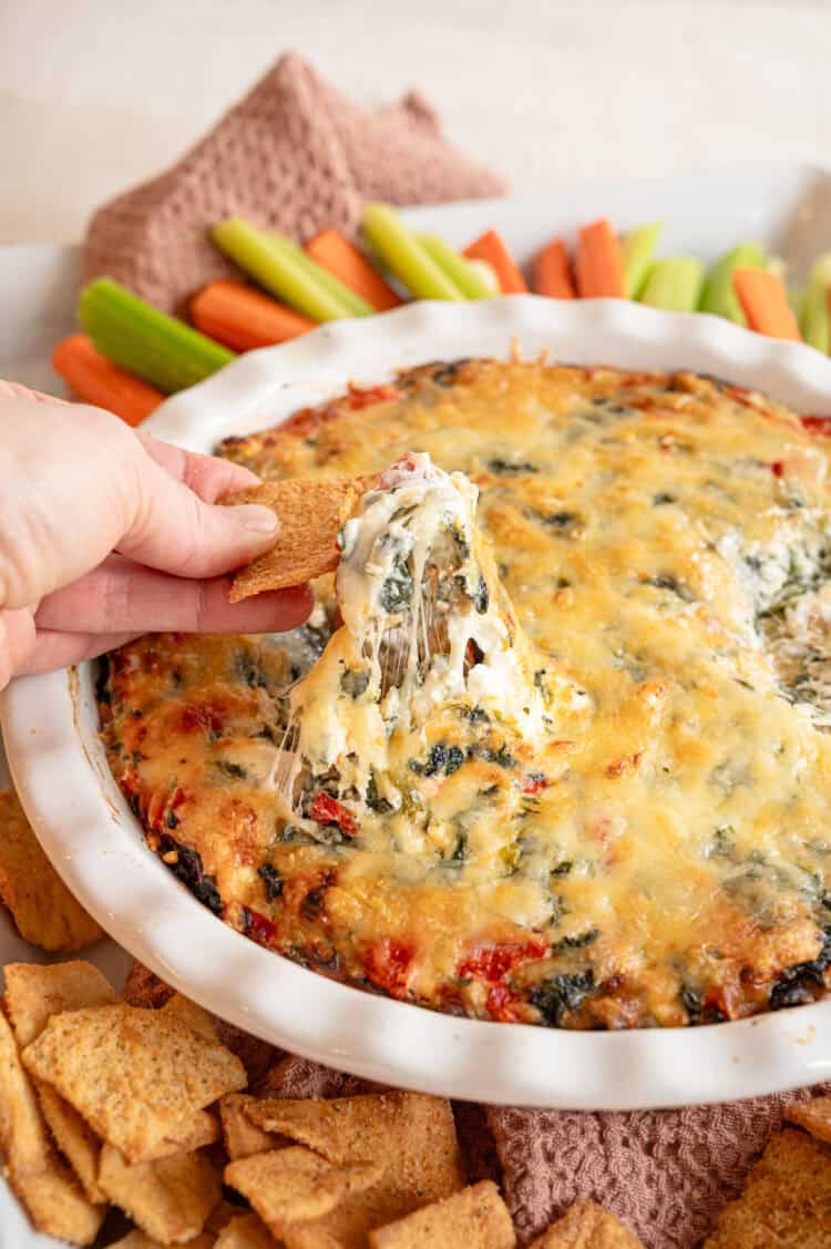 A cracker lifting some spinach and artichoke dip out of the crock it was made in.