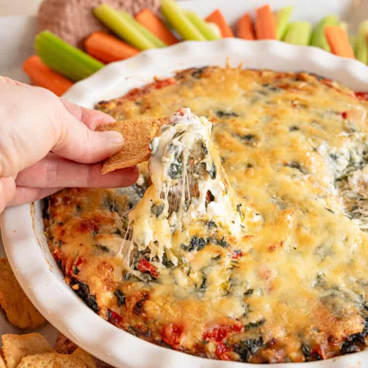 A cracker lifting some spinach and artichoke dip out of the crock it was made in.