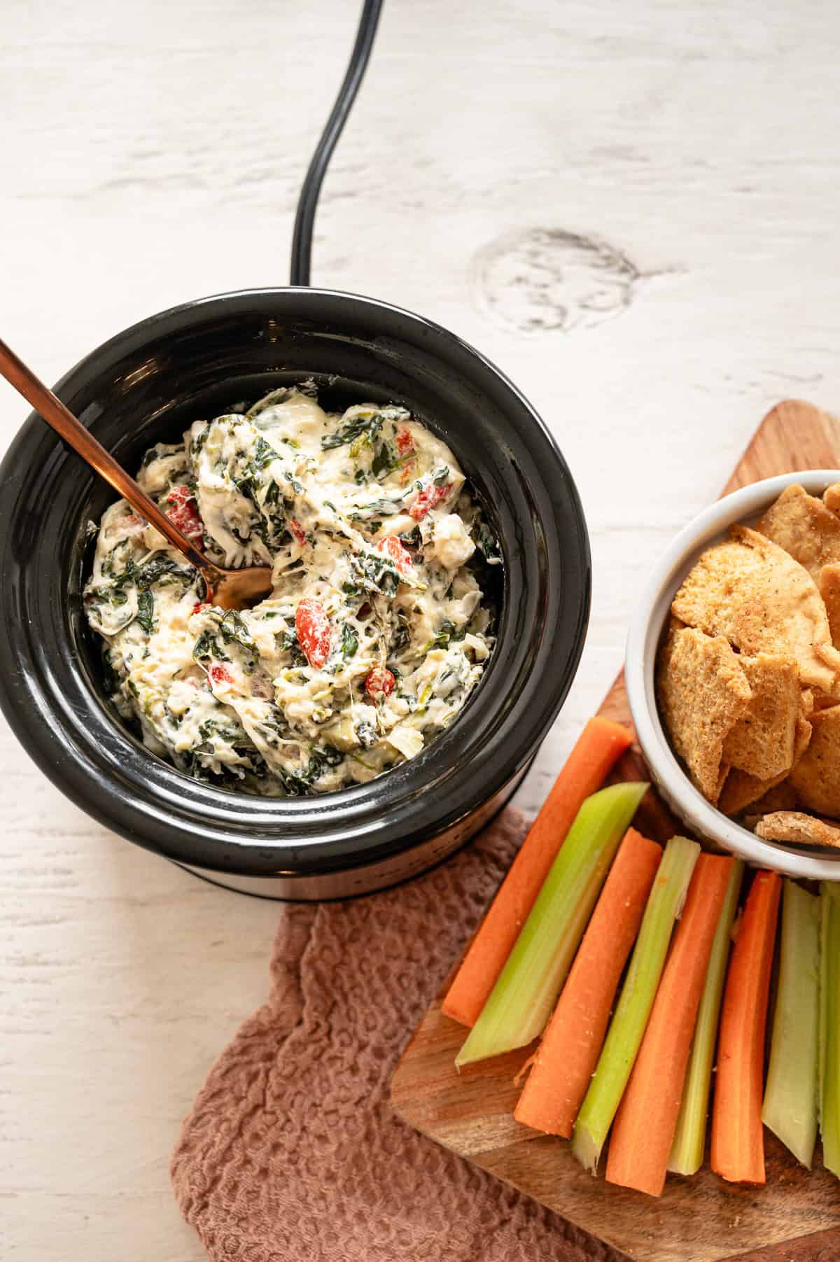 A dipping size crockpot with spinach artichoke dip in it and veggies and crackers on the side.