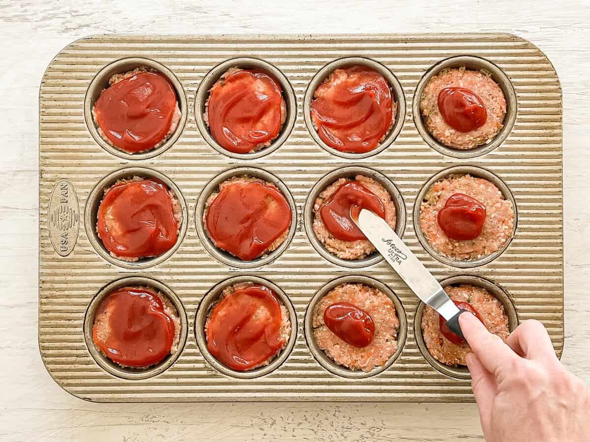 A hand spreading ketchup on top of turkey meatloaf mixture in a muffin tin before baking.