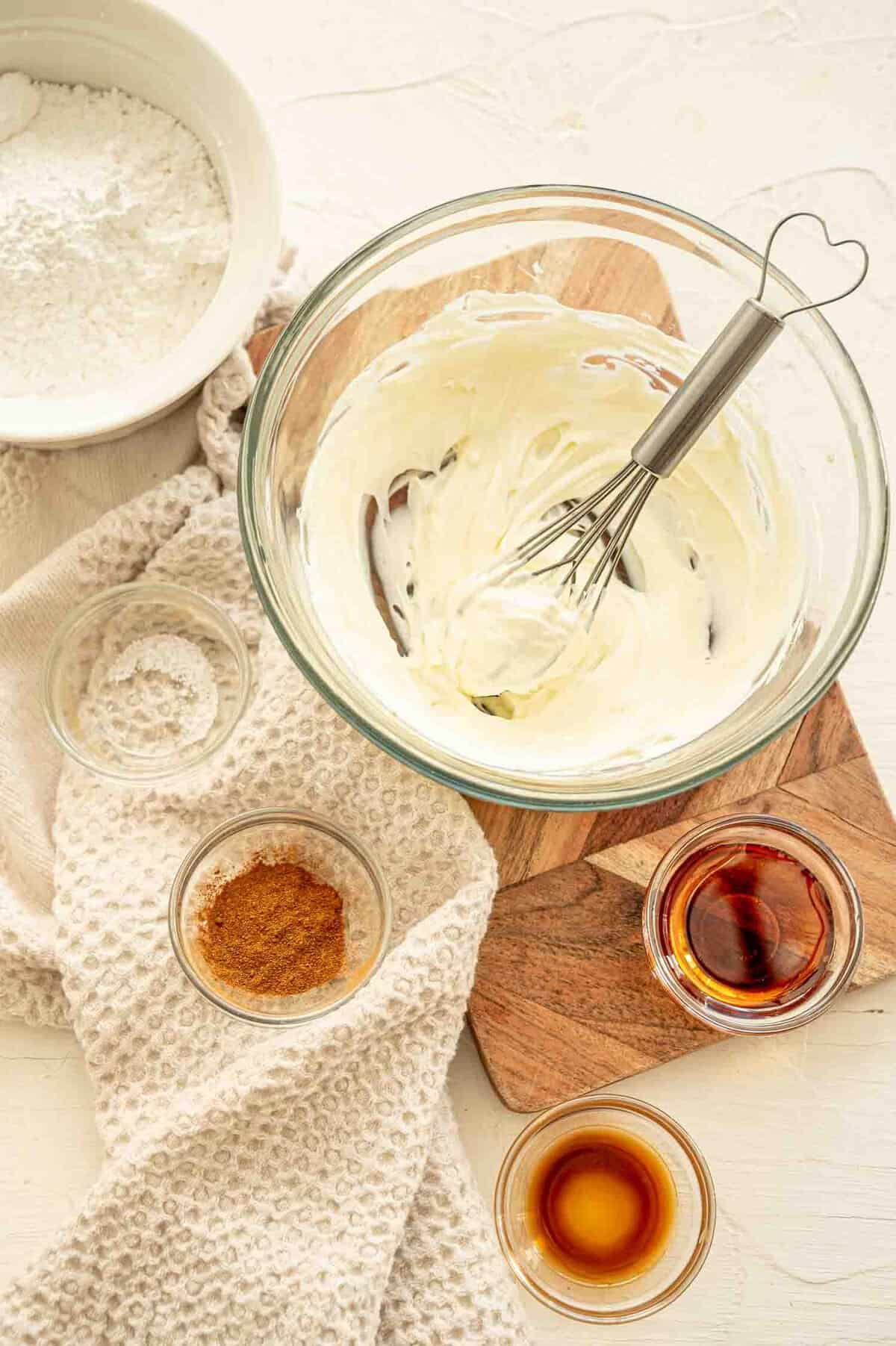 Softened cream cheese in a mixing bowl and smaller bowls of ingredients for cream cheese icing next to it.