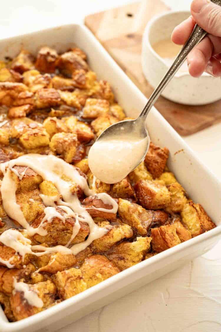 A spoon drizzling cream cheese icing over a 9x12 baking fish of overnight cinnamon roll casserole.