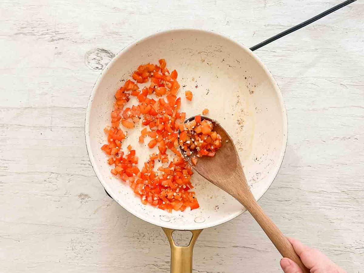 A wooden spoon stirring chopped red pepper and minced garlic in a saute pan.