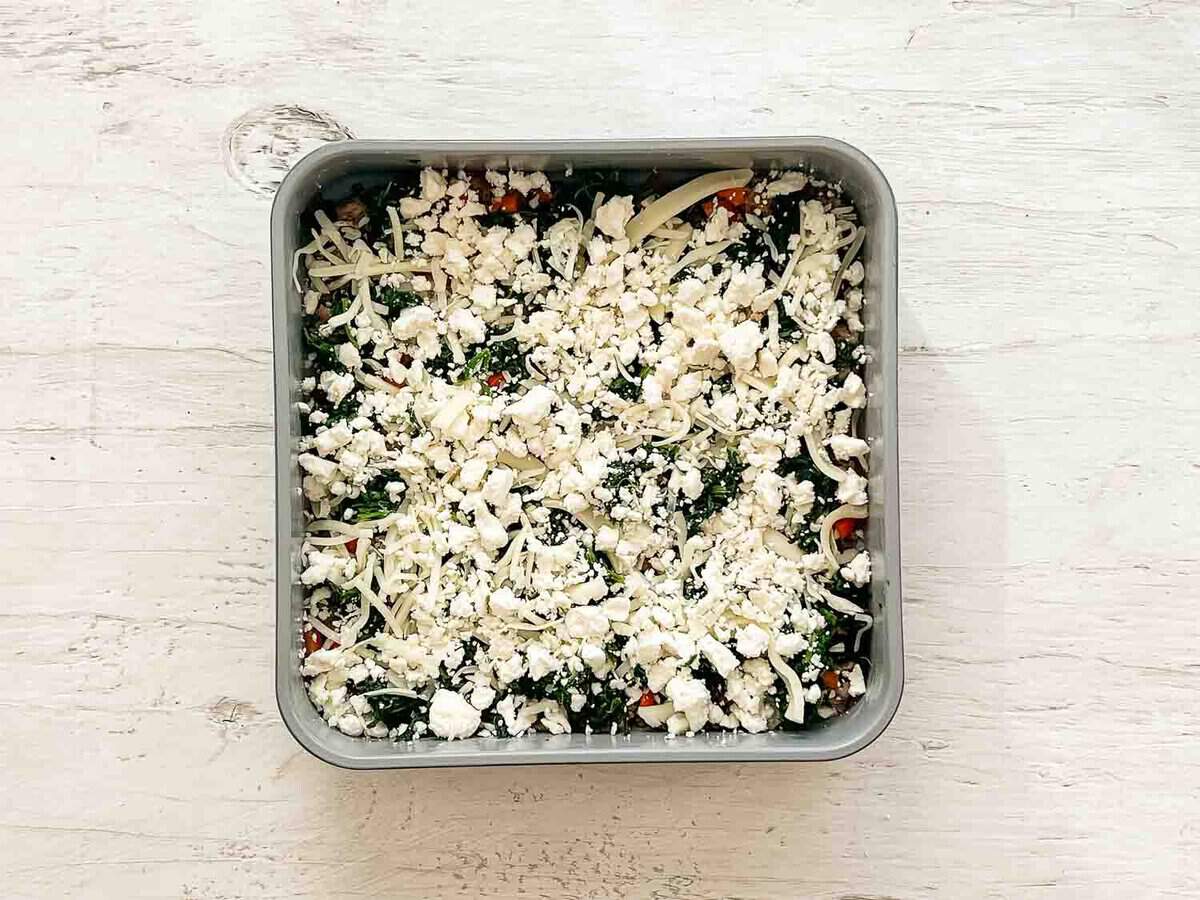 Feta and mozarella cheeses sprinkled on top of sauteed spinach and red peppers, and cooked ground sausage in a baking dish.