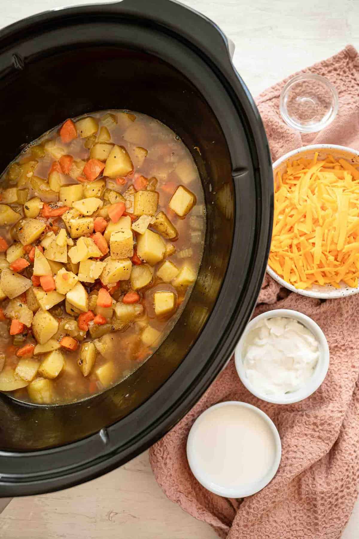 Potato soup ingredients in crockpot with bowls of cheese, half and half, sour cream, and white vinegar next to it.